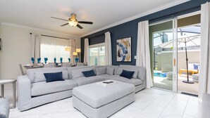 A modern living room with a grey sectional sofa, a small table, and a ceiling fan. - Luxurious Family-friendly 5 Bd 12 Guests w Screened Pool Close to Disney (Kissimmee)