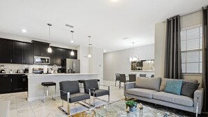 A modern kitchen with dark cabinets, a central island, and pendant lighting. There is a living area with a sofa, chairs, and a glass coffee table. - Four Bedrooms w Pool Townhome 4841 (Kissimmee)