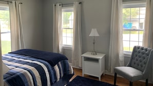 Quarto com cama, mesa de cabeceira, luminária, cadeira e janelas com cortinas.