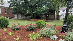Um jardim com diversas plantas e flores, uma cerca de madeira e um edifício de tijolos ao fundo.
