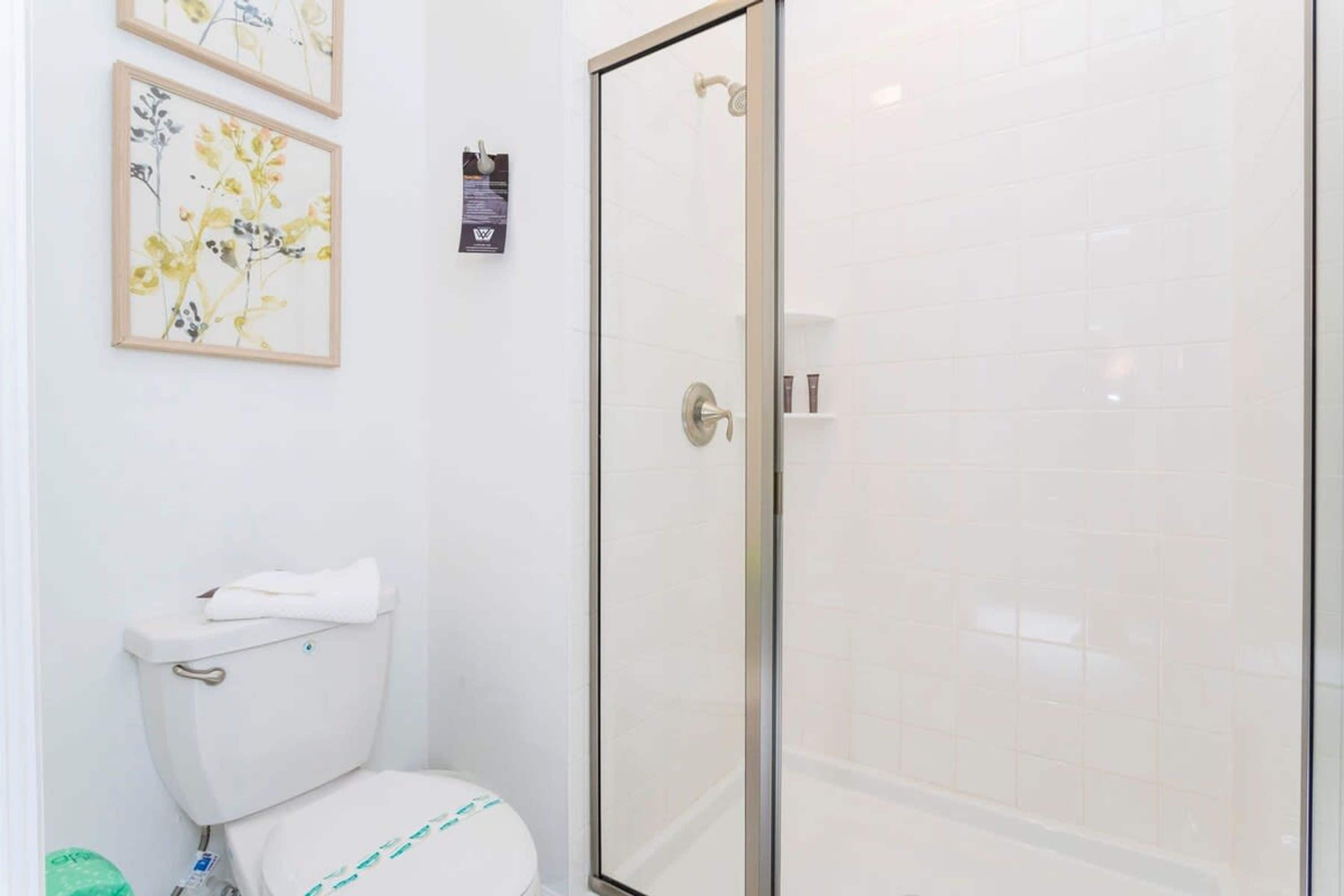 A bathroom with a toilet, a glass shower enclosure, and two framed floral artworks on the wall.