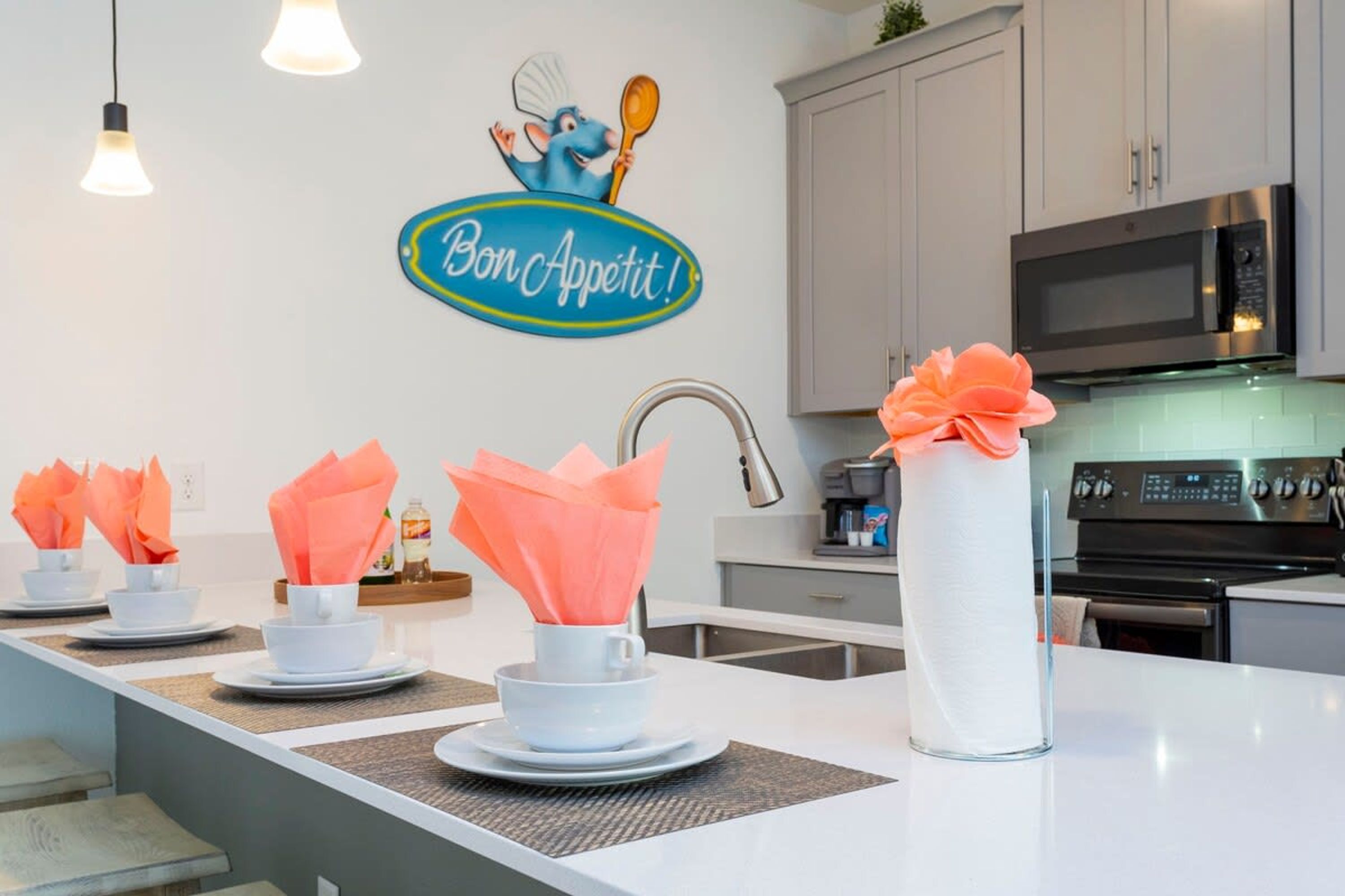 A modern kitchen with white cabinets, a stainless steel microwave, and a sink. There are napkins in white and orange cups on the counter.