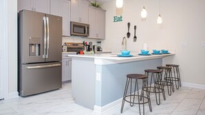 A modern kitchen with a stainless steel refrigerator, a microwave, and a sink. There are bar stools and a blue island. - Wonderful 5 Bedroom w Pool Close to Disney 4801 (Kissimmee)