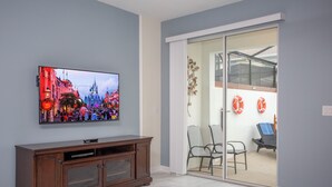 A living room with a flat-screen TV mounted on the wall, a wooden cabinet below it, and a sliding glass door leading to an outdoor seating area with chairs and a table. - Wonderful 5 Bedroom w Pool Close to Disney 4801 (Kissimmee)