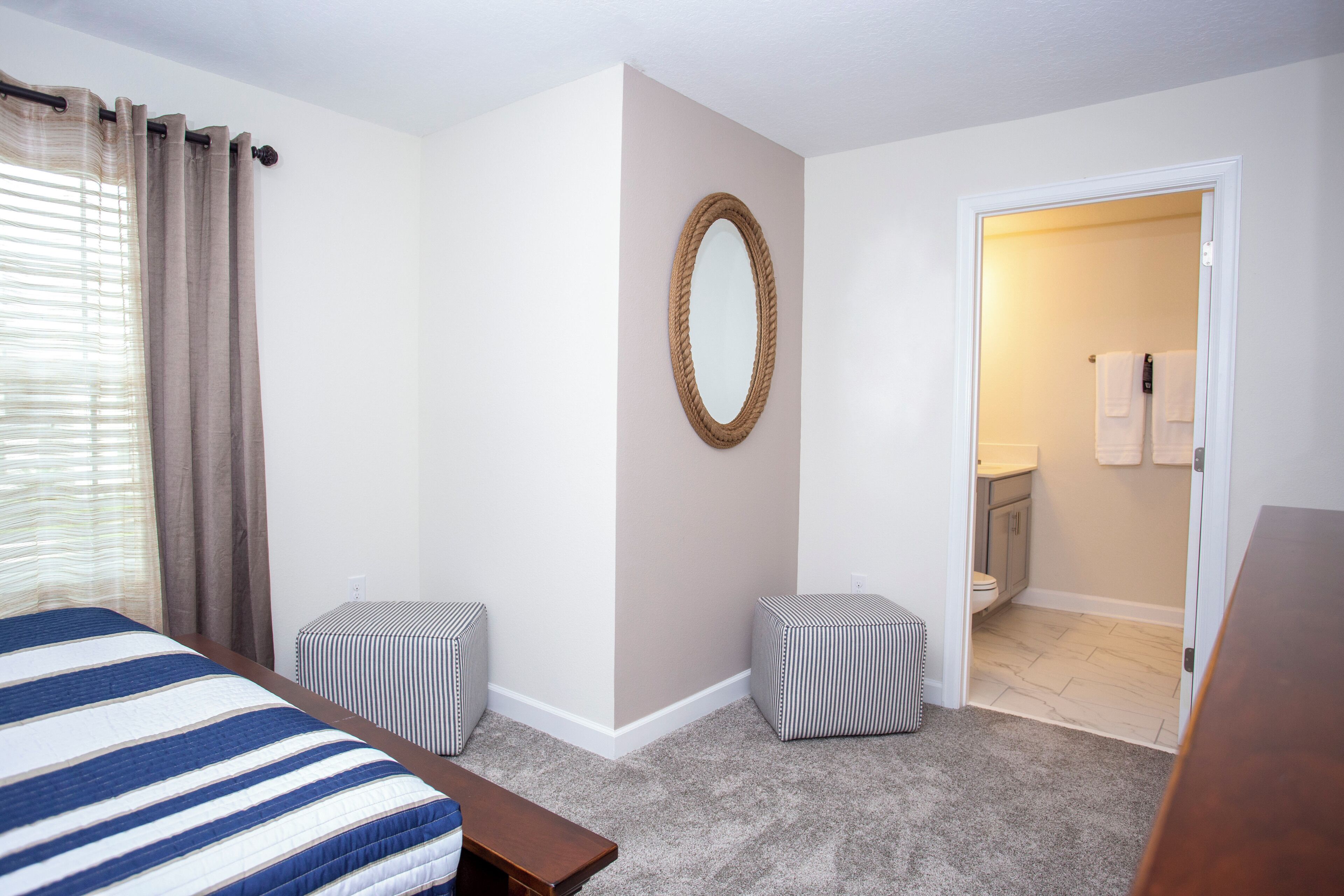 Una camera da letto con un letto, due poggiapiedi, uno specchio e un bagno parzialmente visibile attraverso una porta aperta.
