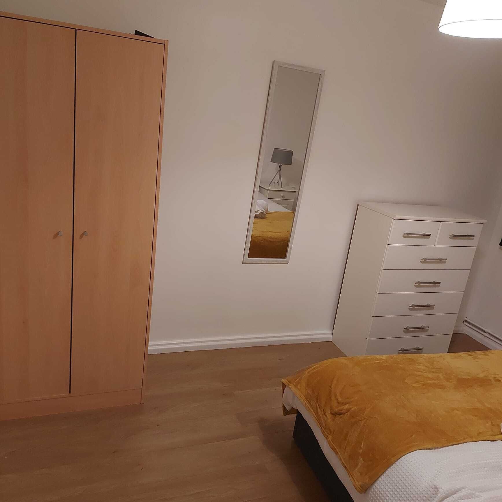 Un dormitorio con un armario de madera, un tocador blanco, una cama con una colcha amarilla y un espejo en la pared.