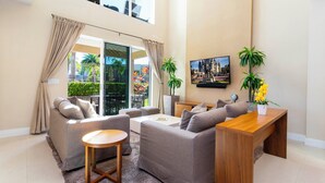 A modern living room with a grey sofa set, a wooden coffee table, and a wooden side table. There is a large window with curtains, a painting on the wall, and a potted plant. - Marvelous 4 Bd Close to Disney Magic Village 3153 (Kissimmee)