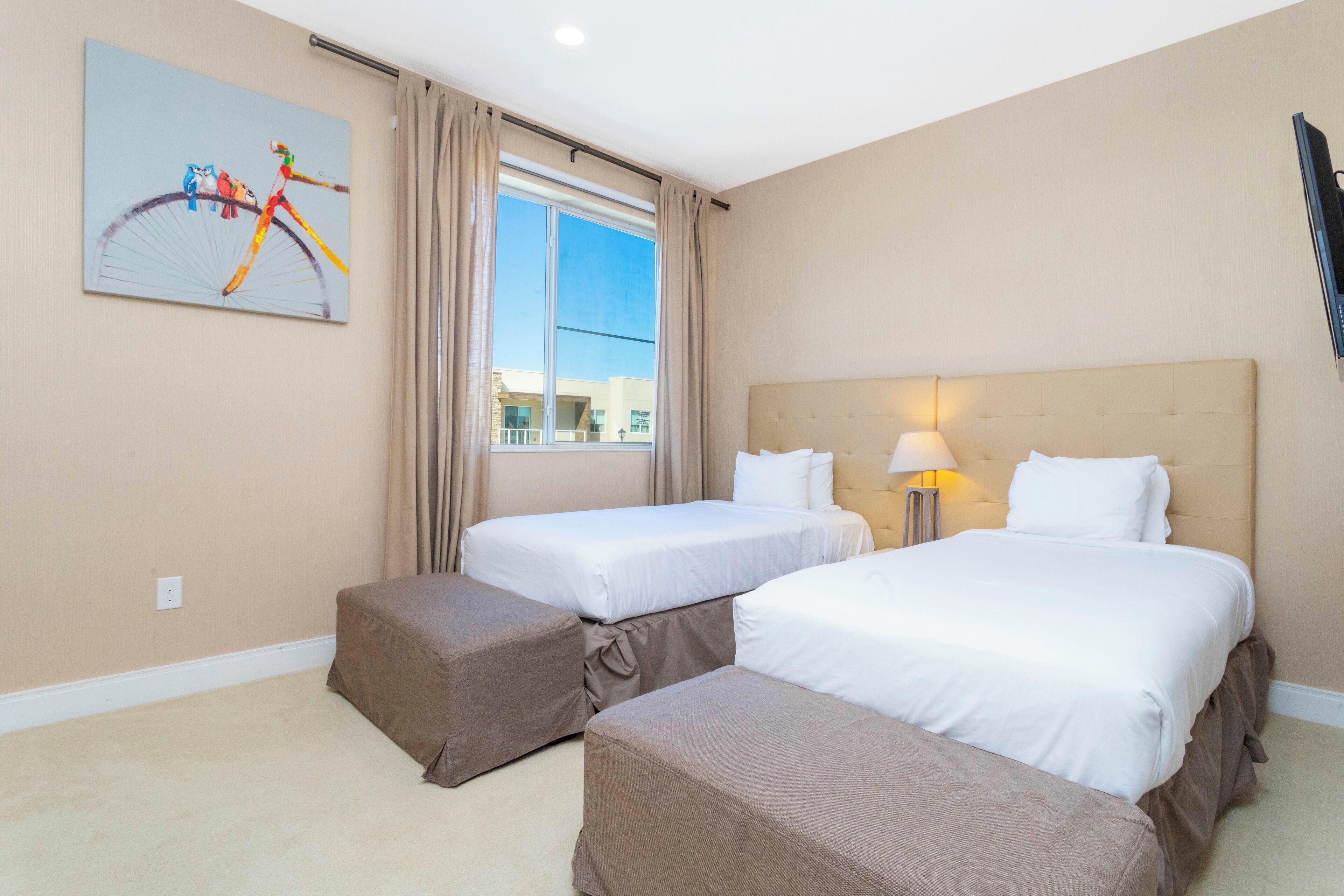 A hotel room with two beds, a large window with curtains, a painting on the wall, and a flat-screen TV mounted on the wall.
