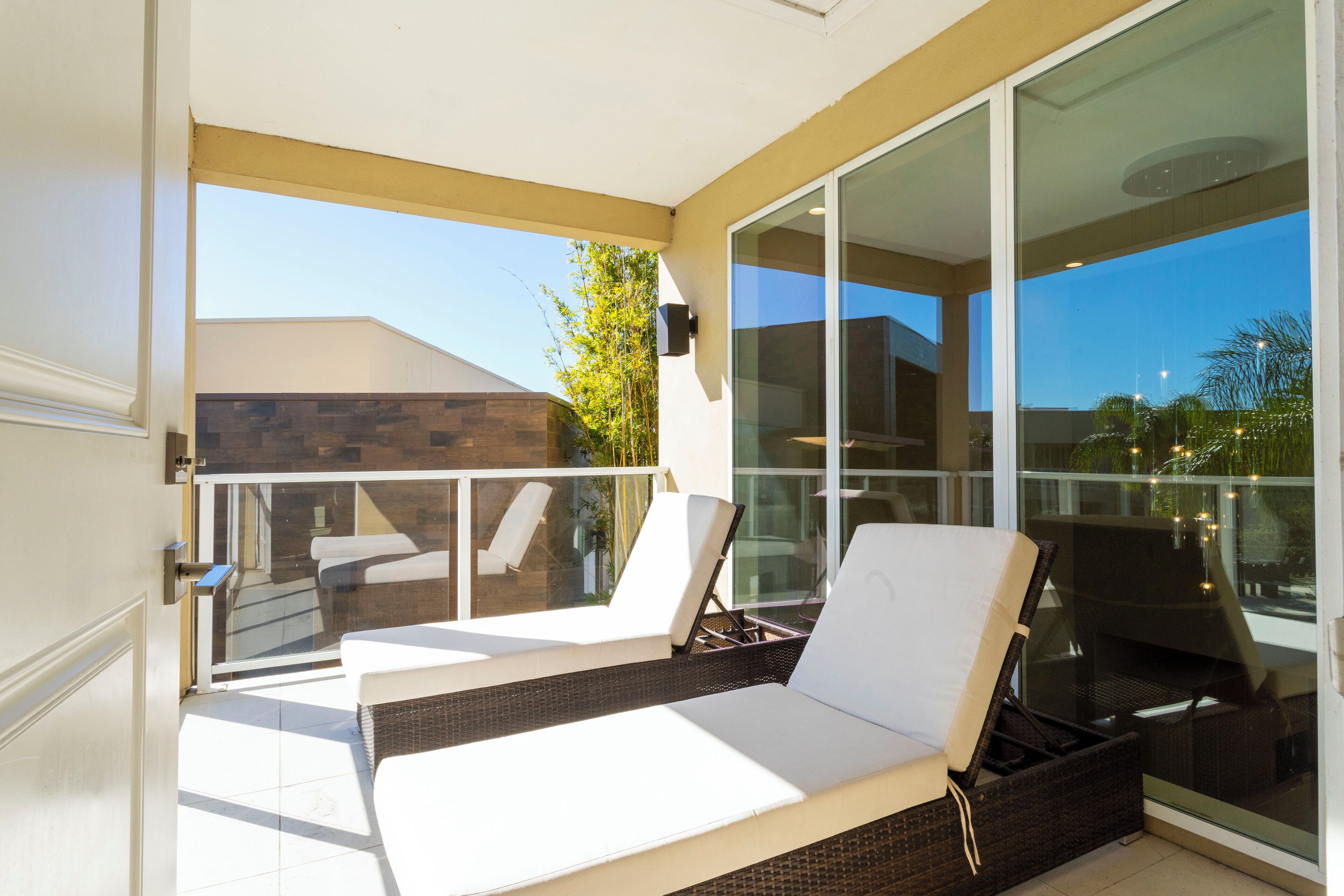 Un balcon avec deux fauteuils de jardin blancs, une table en osier et une porte vitrée donnant sur un espace patio.
