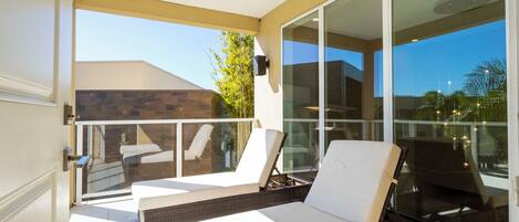 Un balcon avec deux fauteuils de jardin blancs, une table en osier et une porte vitrée donnant sur un espace patio.