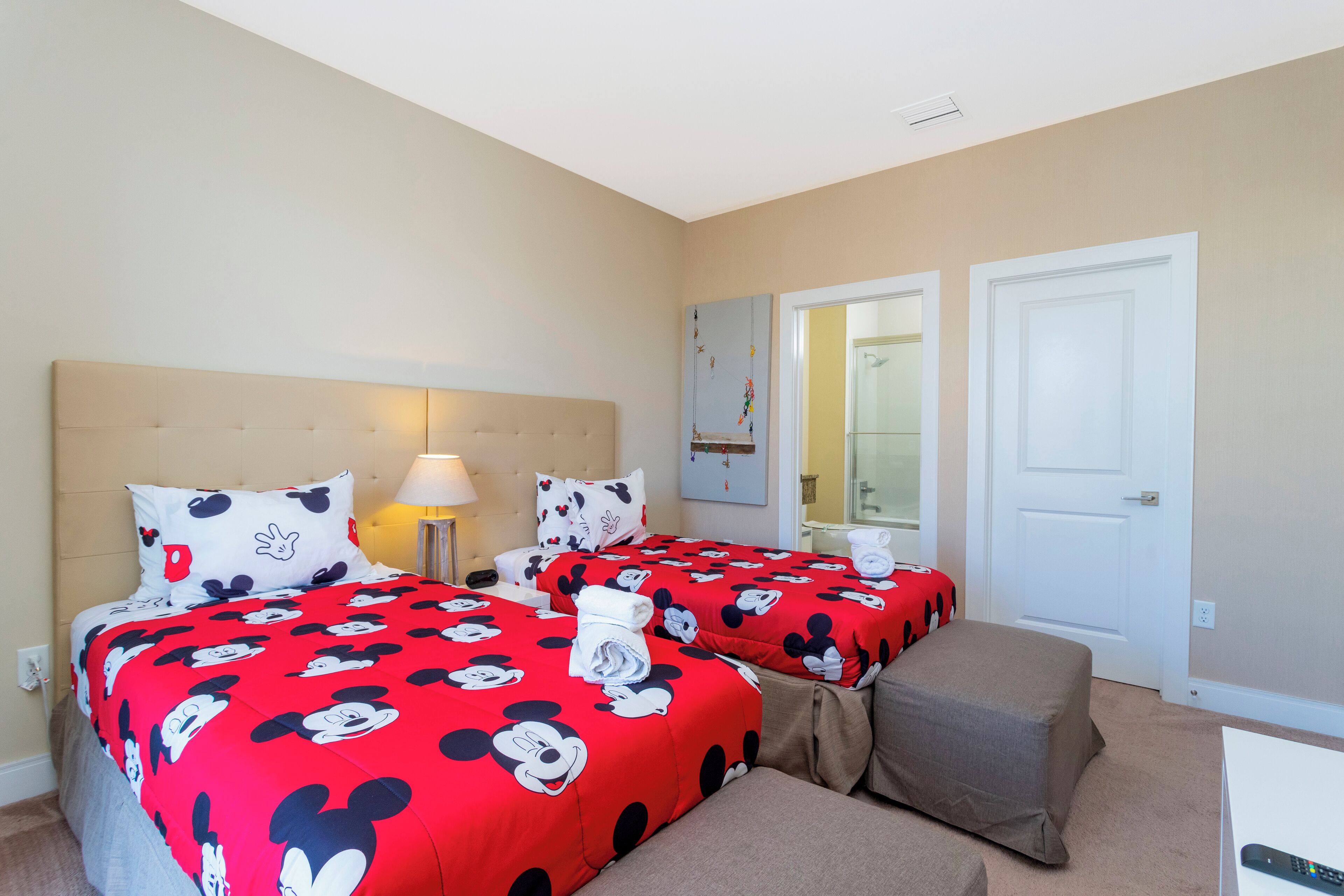 Et hotelværelse med to senge, begge med sengetøj med Mickey Mouse tema, et natbord med lampe, en lille bænk og en dør til badeværelset.