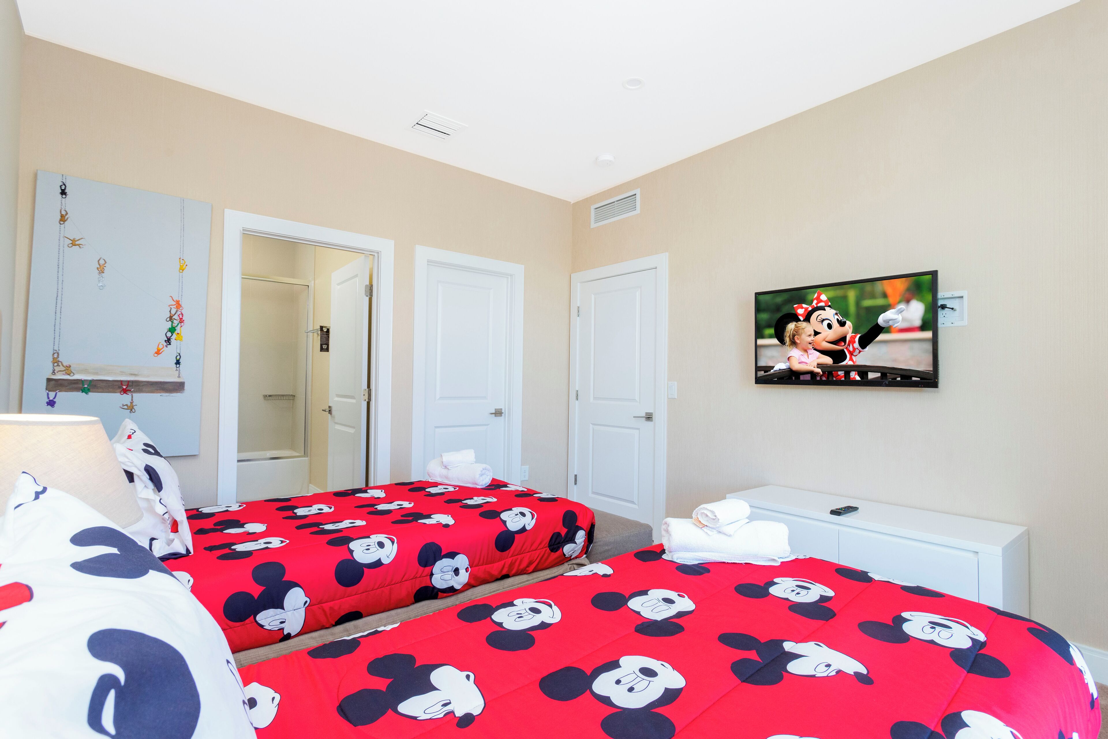 Ein Hotelzimmer mit zwei Betten, jeweils mit einer roten Bettdecke im Mickey-Mouse-Design, einem weißen Nachttisch und einem an der Wand befestigten Fernseher.