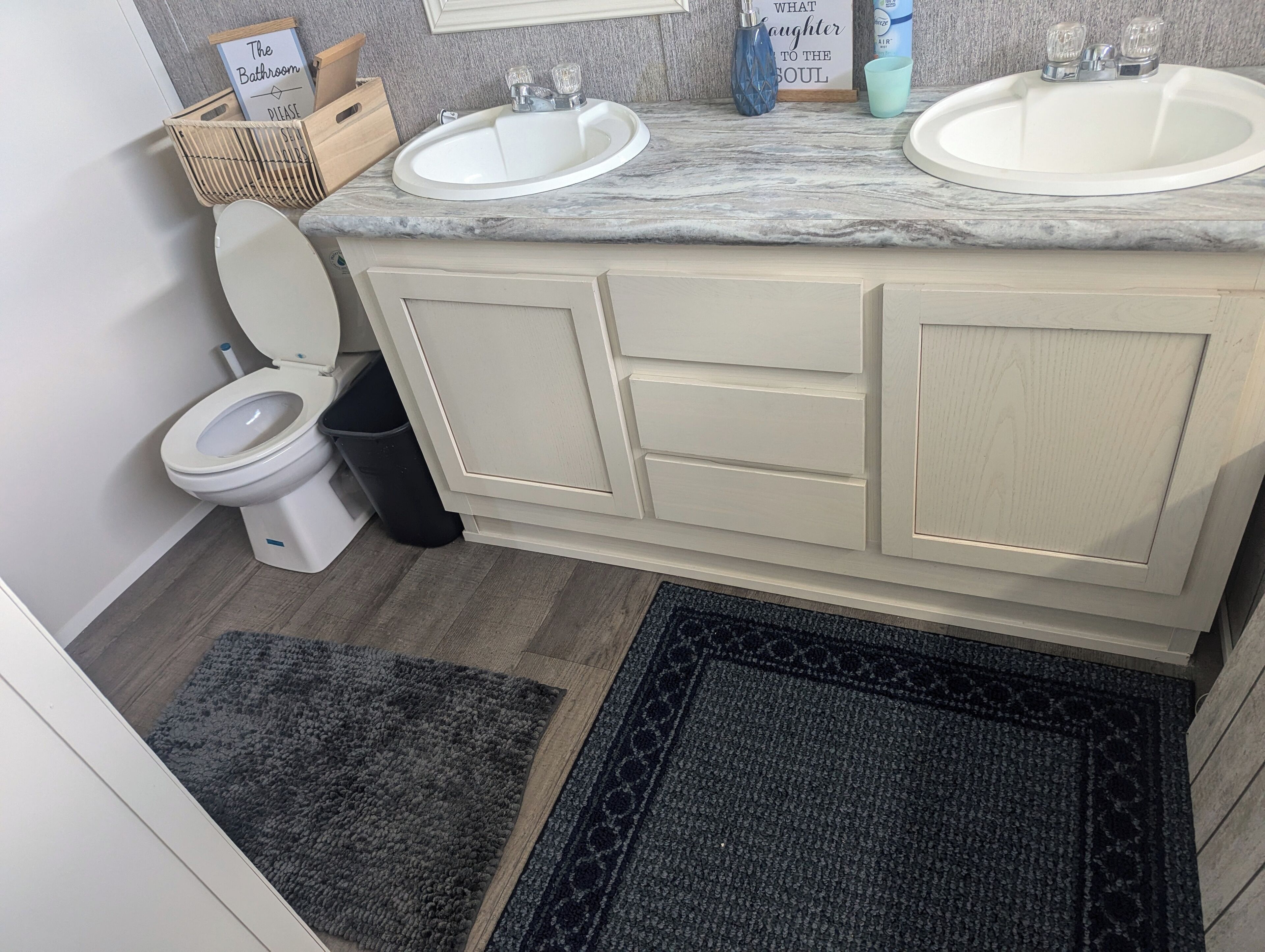 A bathroom with two sinks, a toilet, and a trash can. There are two rugs on the floor.