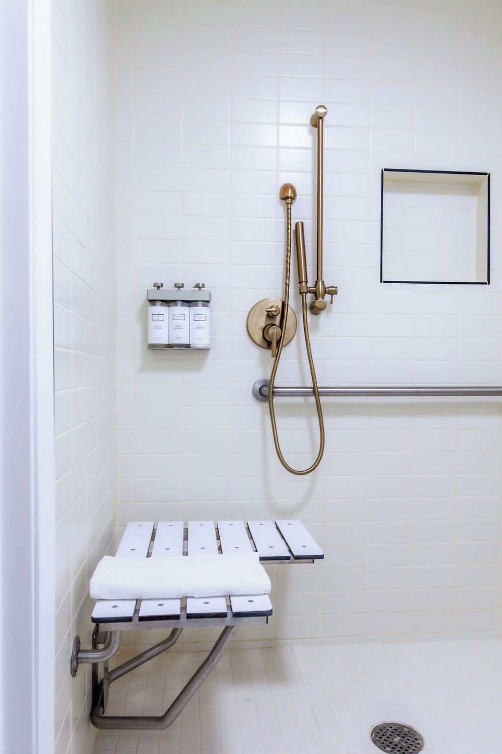 A shower area with a handheld showerhead, a wall-mounted faucet, and a grab bar. There are two bottles of shampoo and conditioner on a shelf.