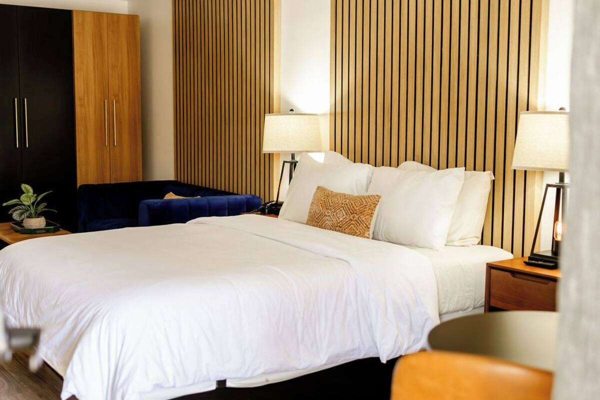 A hotel room with a large bed, two bedside lamps, a wooden panel wall, a dark blue chair, and a small plant on a side table.