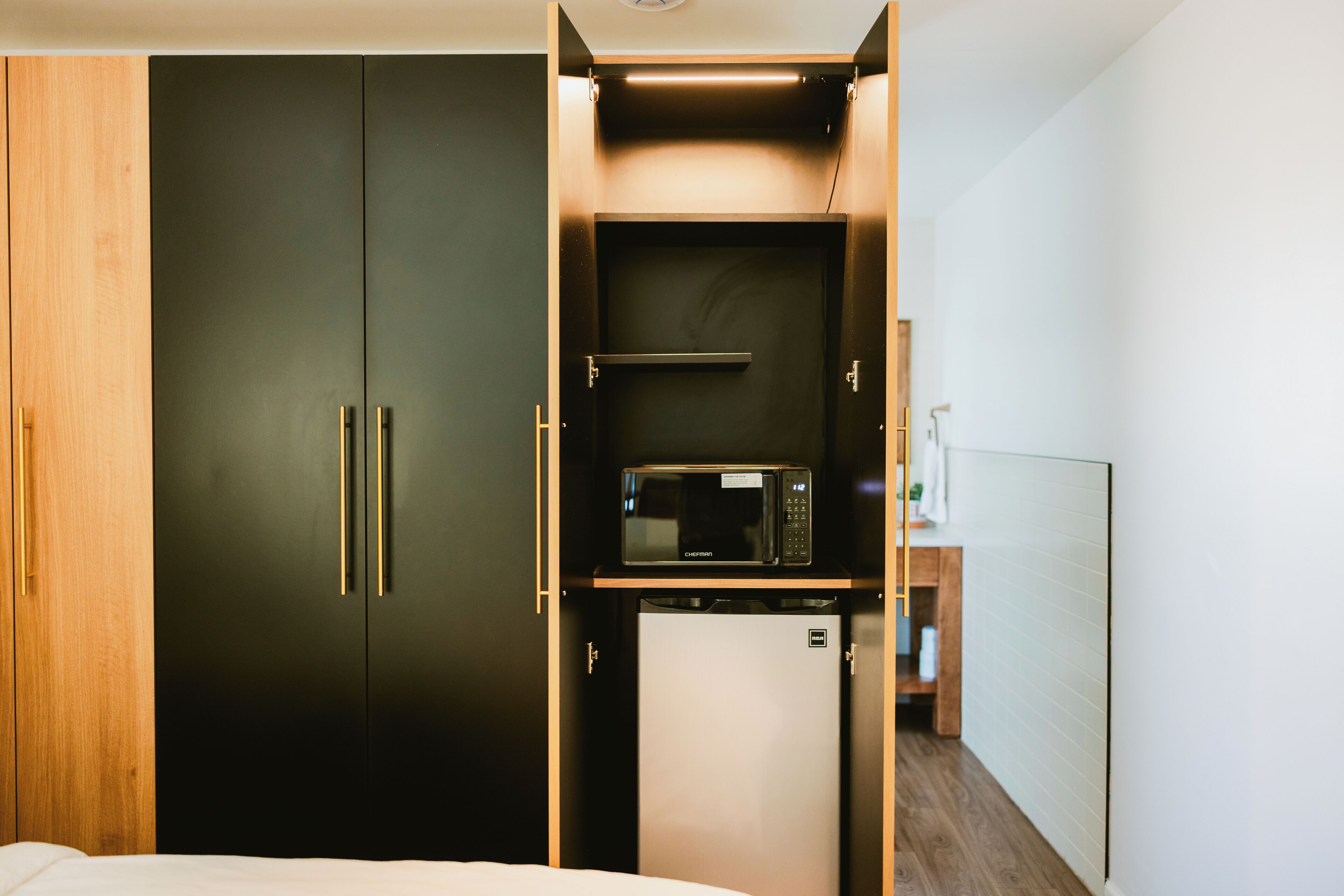 A compact room with a bed, a dark wardrobe, a small kitchenette with a microwave, and a whiteboard.