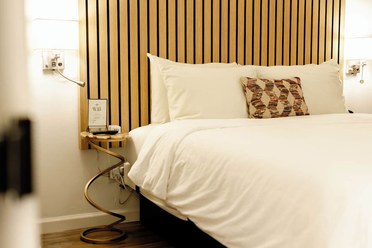 A neatly made bed with white linens, a wooden slat headboard, and a bedside table with a lamp and a power outlet.