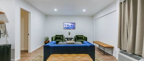 A modern living room with a blue sofa, a wooden bench, a TV mounted on the wall, and a large window with curtains.