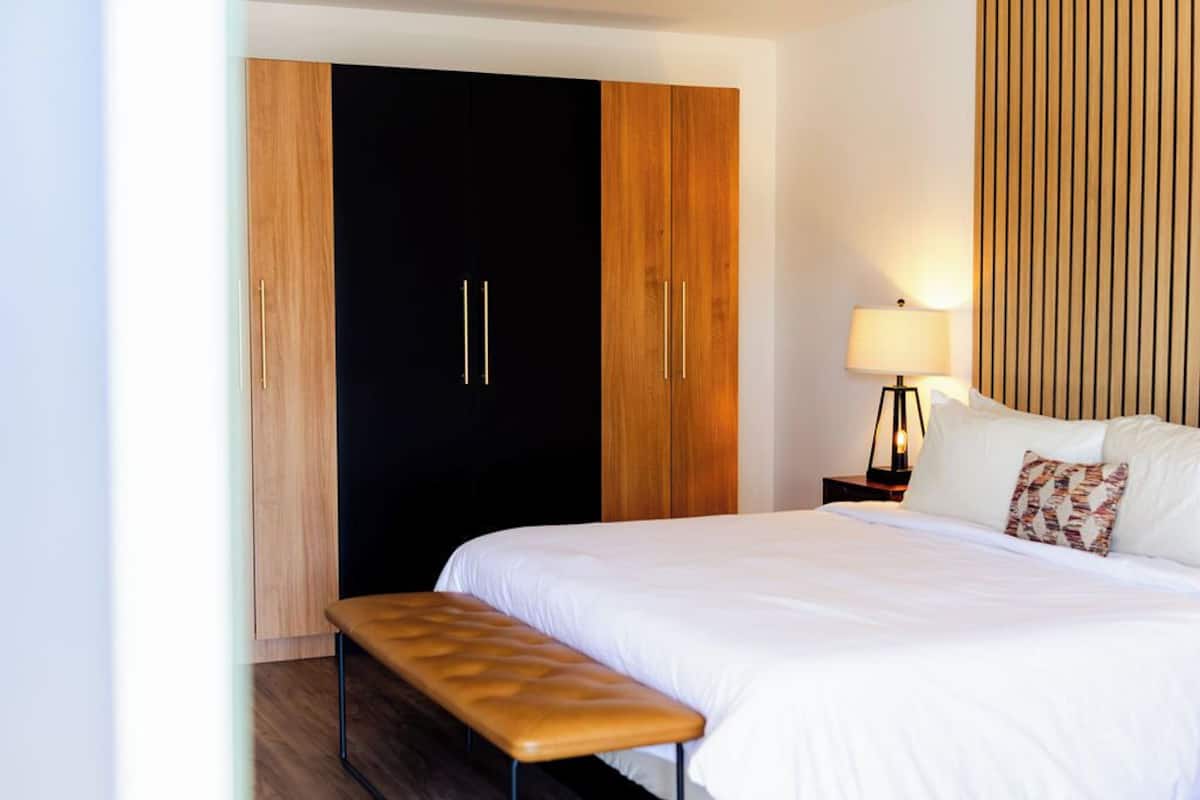 A hotel room with a bed, a bench, a wardrobe, and a nightstand with a lamp.