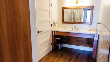 Un baño con suelo de madera, paredes blancas, una puerta blanca y un tocador de madera con un espejo, además de un lavabo blanco.