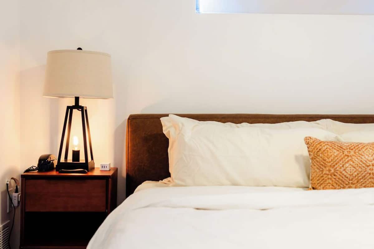A neatly made bed with white linens and a patterned pillow, a bedside lamp, and a wooden nightstand with a phone and a power outlet.