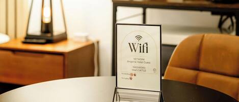 A hotel room with a round table, a lamp, a sign indicating WiFi availability, and a coffee machine.