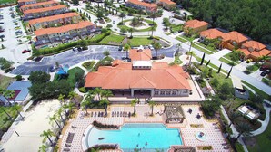 An aerial view of a resort with a central swimming pool, multiple buildings with red roofs, and a parking area.