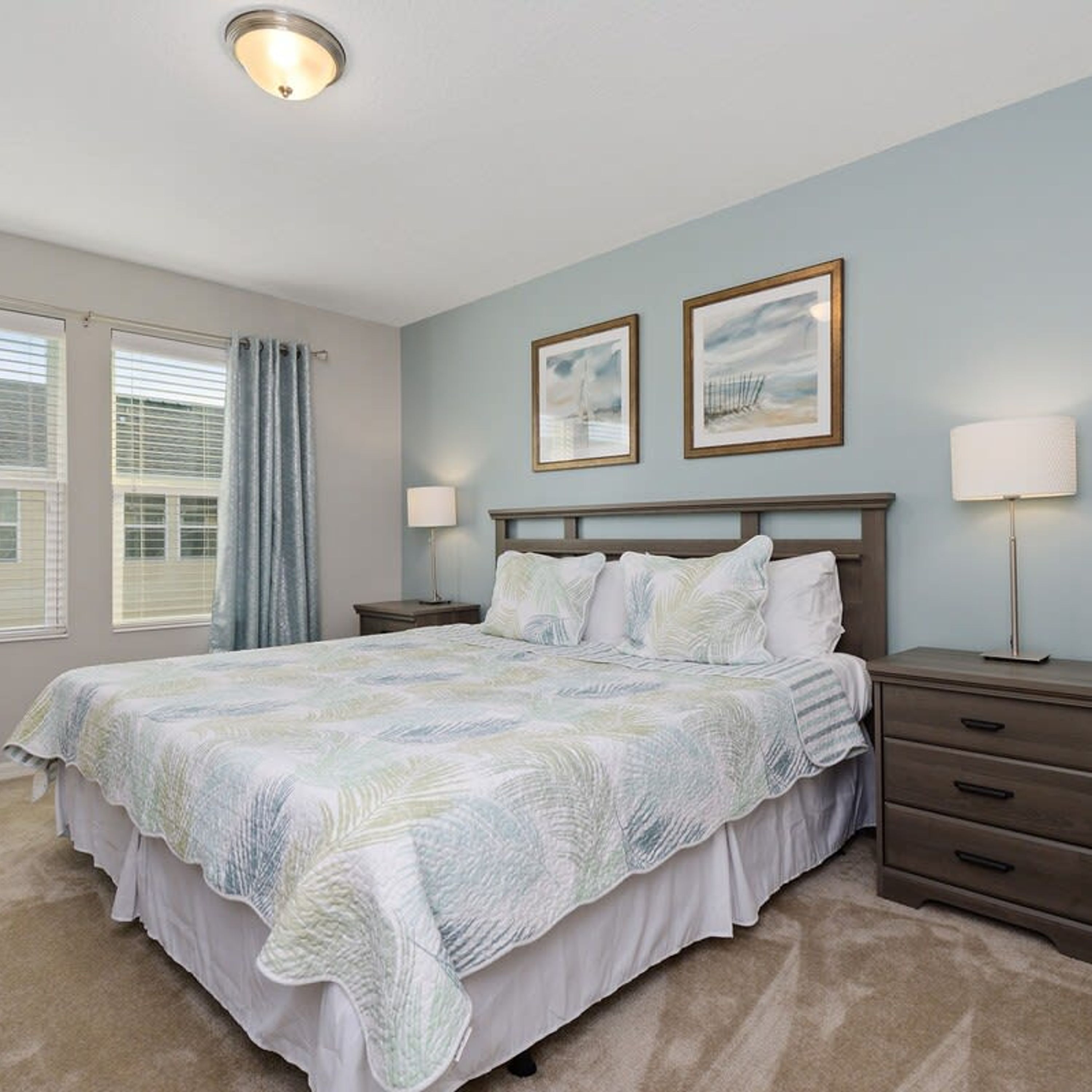 A bedroom with a bed, two bedside tables, a nightstand, a ceiling light, and two framed pictures on the wall.
