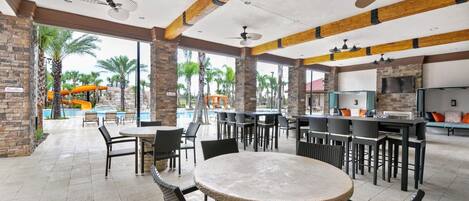 A covered outdoor dining area with tables and chairs, a pool with slides, and a bar area with seating.
