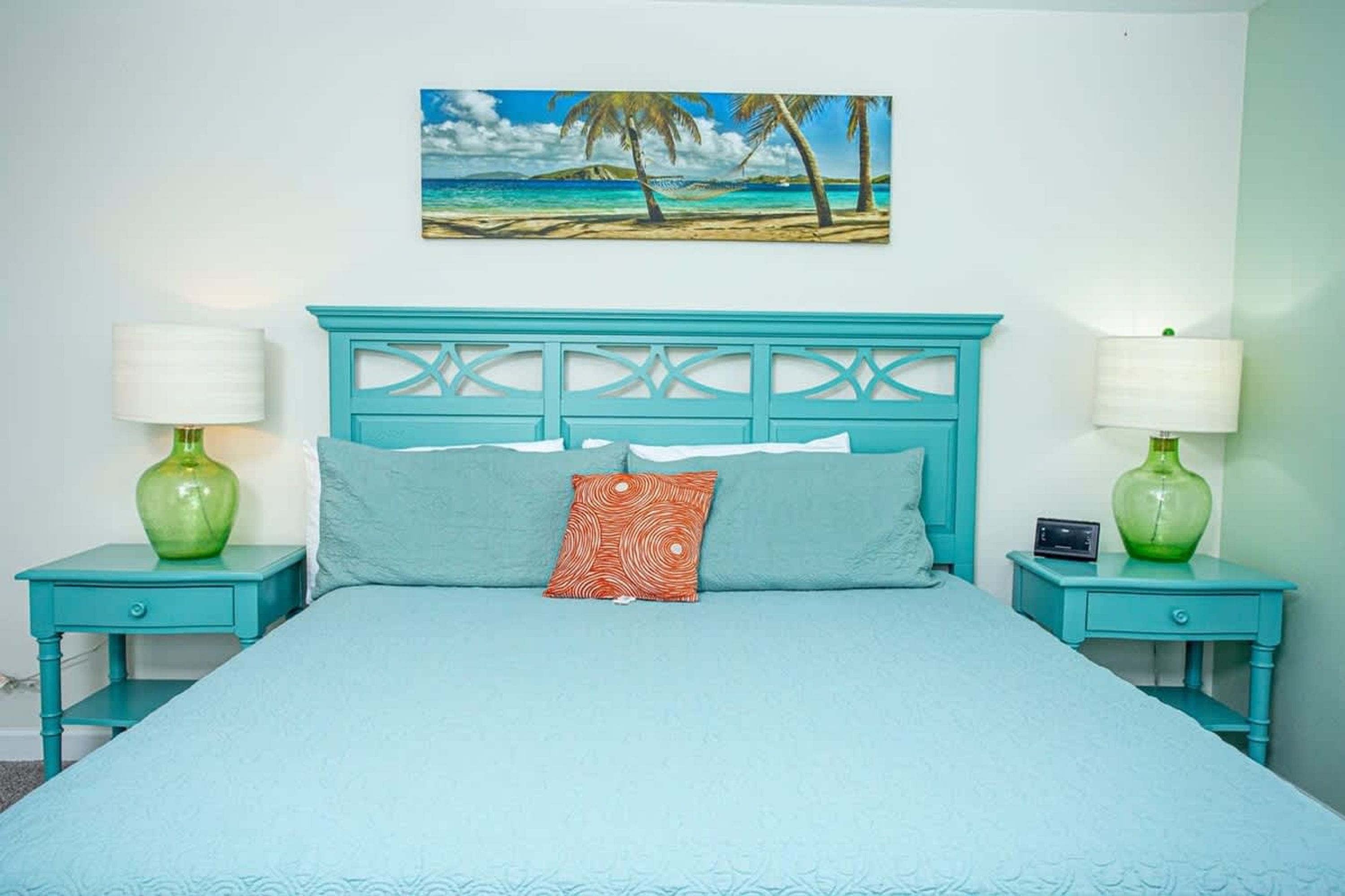 A bed with a turquoise headboard, two nightstands with lamps, and a painting of a beach scene above the bed.
