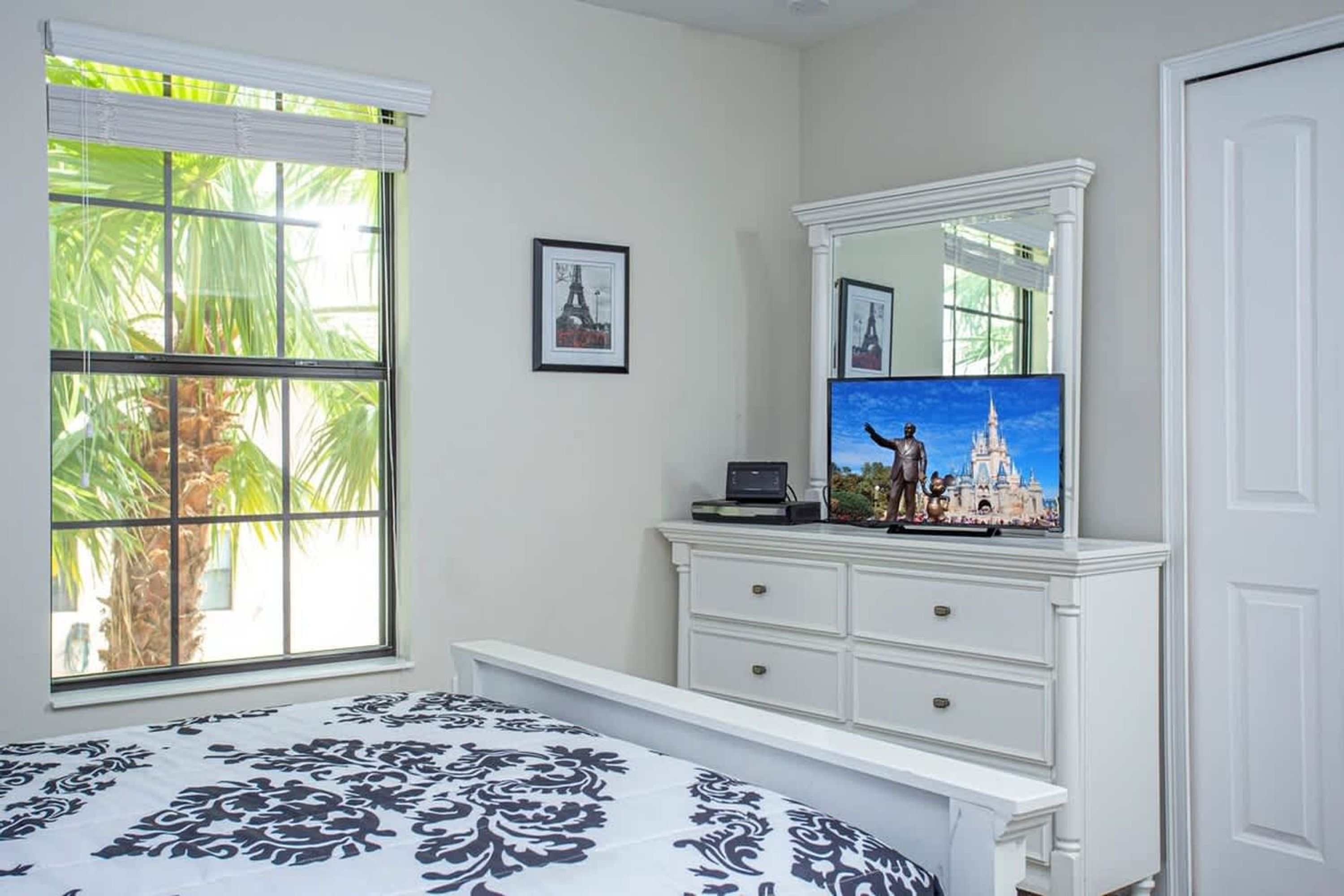 A bedroom with a bed, a dresser, a mirror, and a window with a view of palm trees.