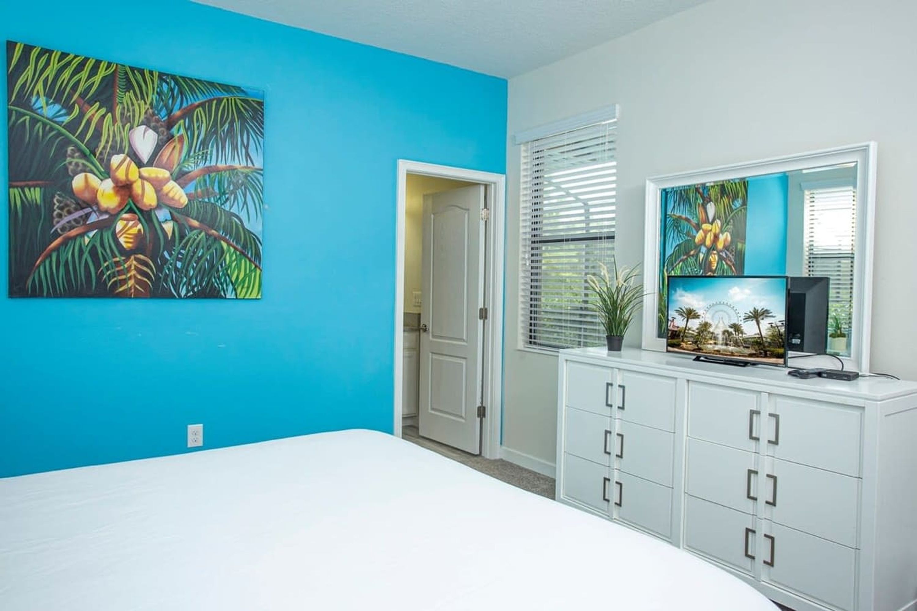 Ein Schlafzimmer mit blauer Wand, einem weißen Bett und einer weißen Kommode mit Fernseher, sowie einer Tür zu einem weiteren Raum.