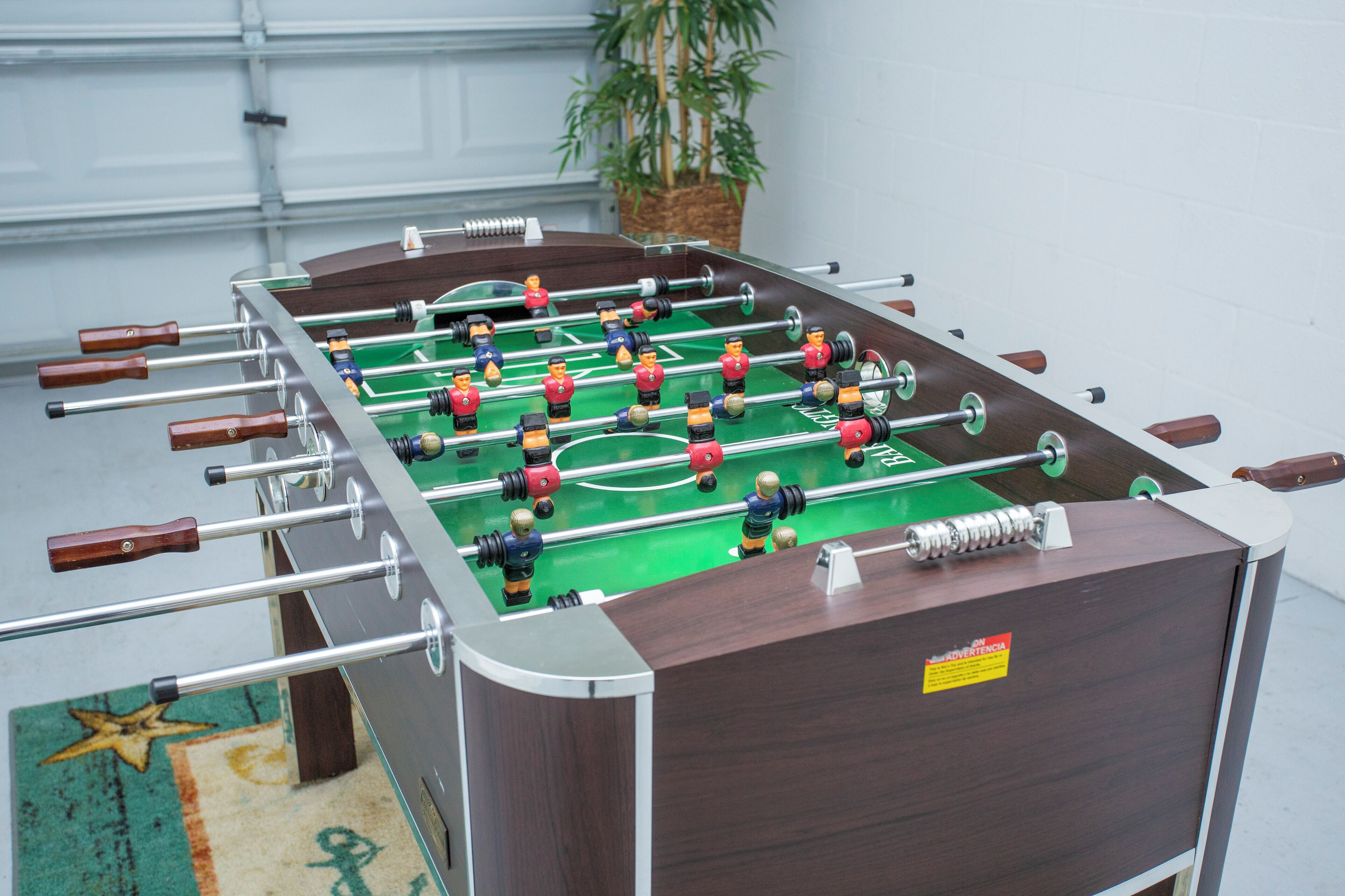 Ein Kicker mit mehreren Spielern und Stangen vor einer weißen Wand, im Hintergrund ist eine Pflanze zu sehen.
