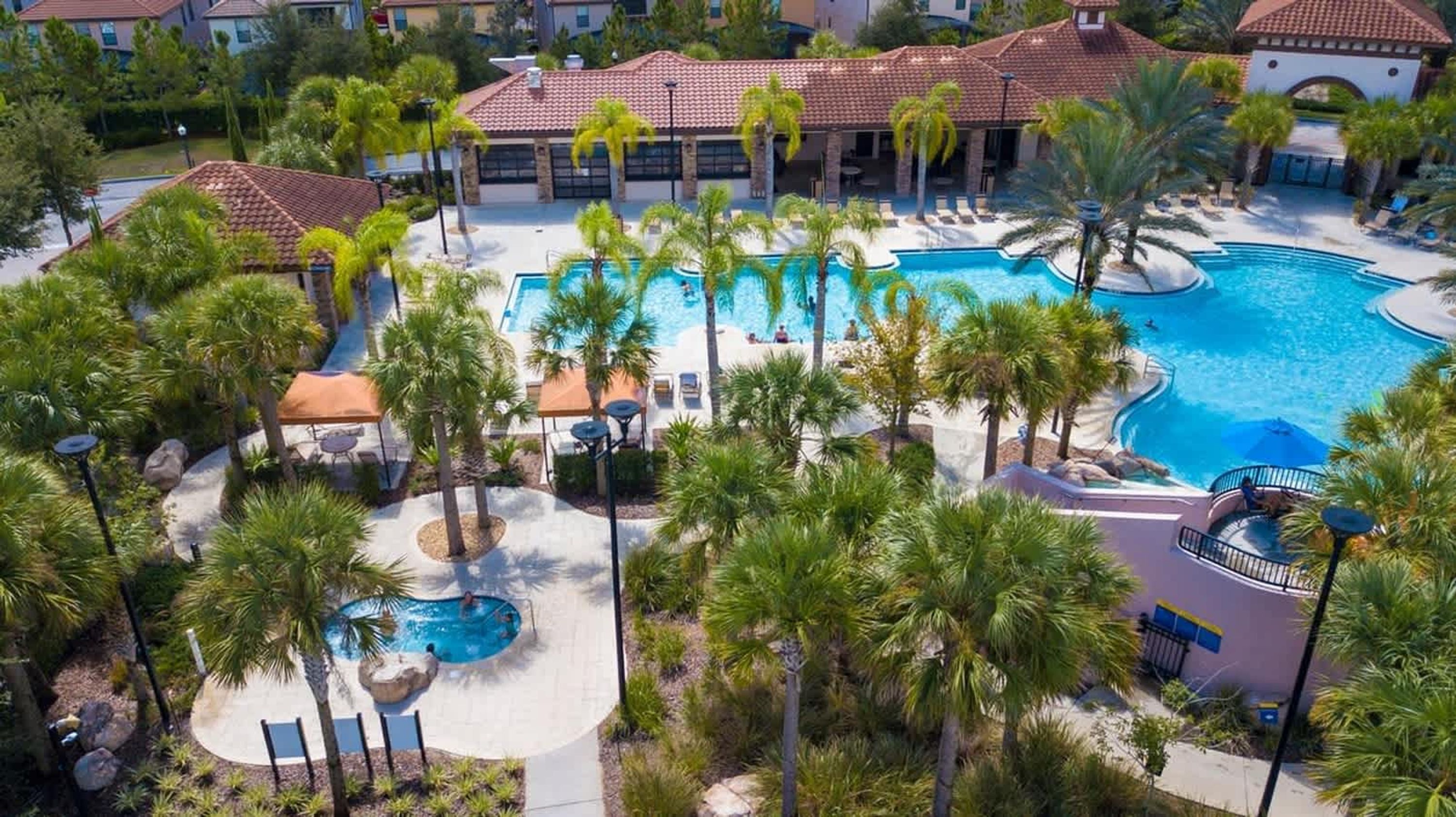A resort with a large central pool, palm trees, and a building with red-tiled roofs.