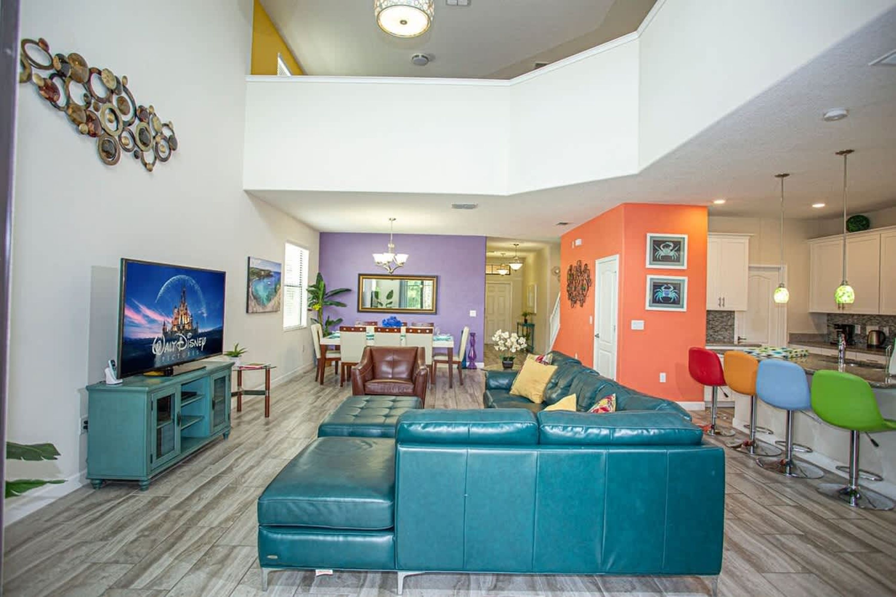 Un salon spacieux avec un grand canapé de couleur turquoise, un téléviseur et un coin repas agrémenté d’un mur violet et d’un lustre.