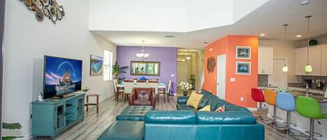 Un salon spacieux avec un grand canapé de couleur turquoise, un téléviseur et un coin repas agrémenté d’un mur violet et d’un lustre.
