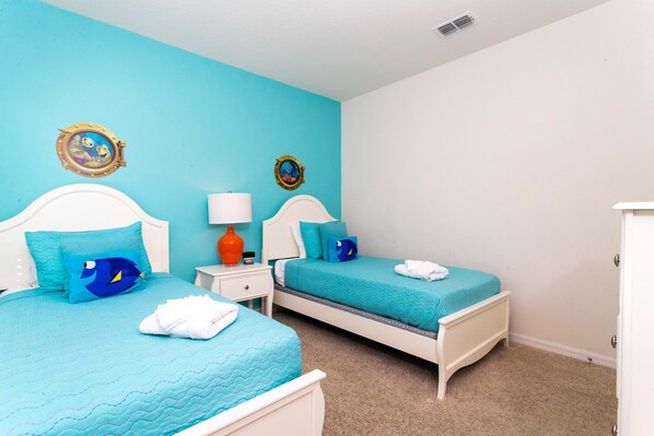 Una habitación con dos camas, cada una con una colcha azul y almohadas a juego. Hay dos adornos en la pared, un espejo redondo y una mesita de noche con una lámpara.