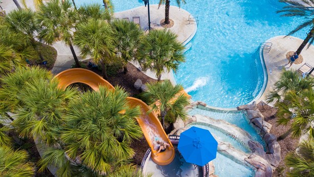 Luchtbeeld van een zwembadgedeelte van een resort, met een glijbaan, palmbomen en een rond zwembad.