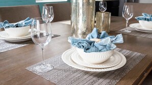 Een eettafel gedekt met witte borden, kommen en wijnglazen, met een blauwe tafelkleed en een broodmand.