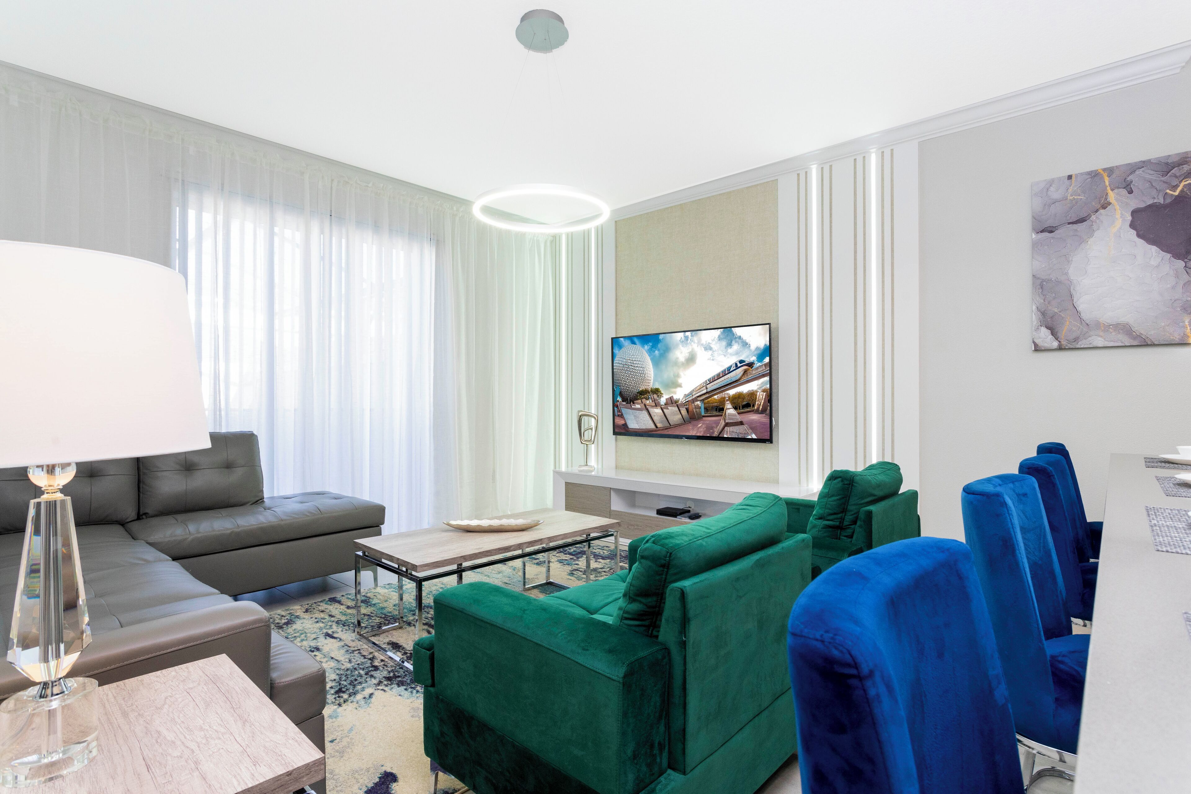 A modern hotel room with a grey sofa, a green sofa, a dining table, and a TV mounted on the wall.