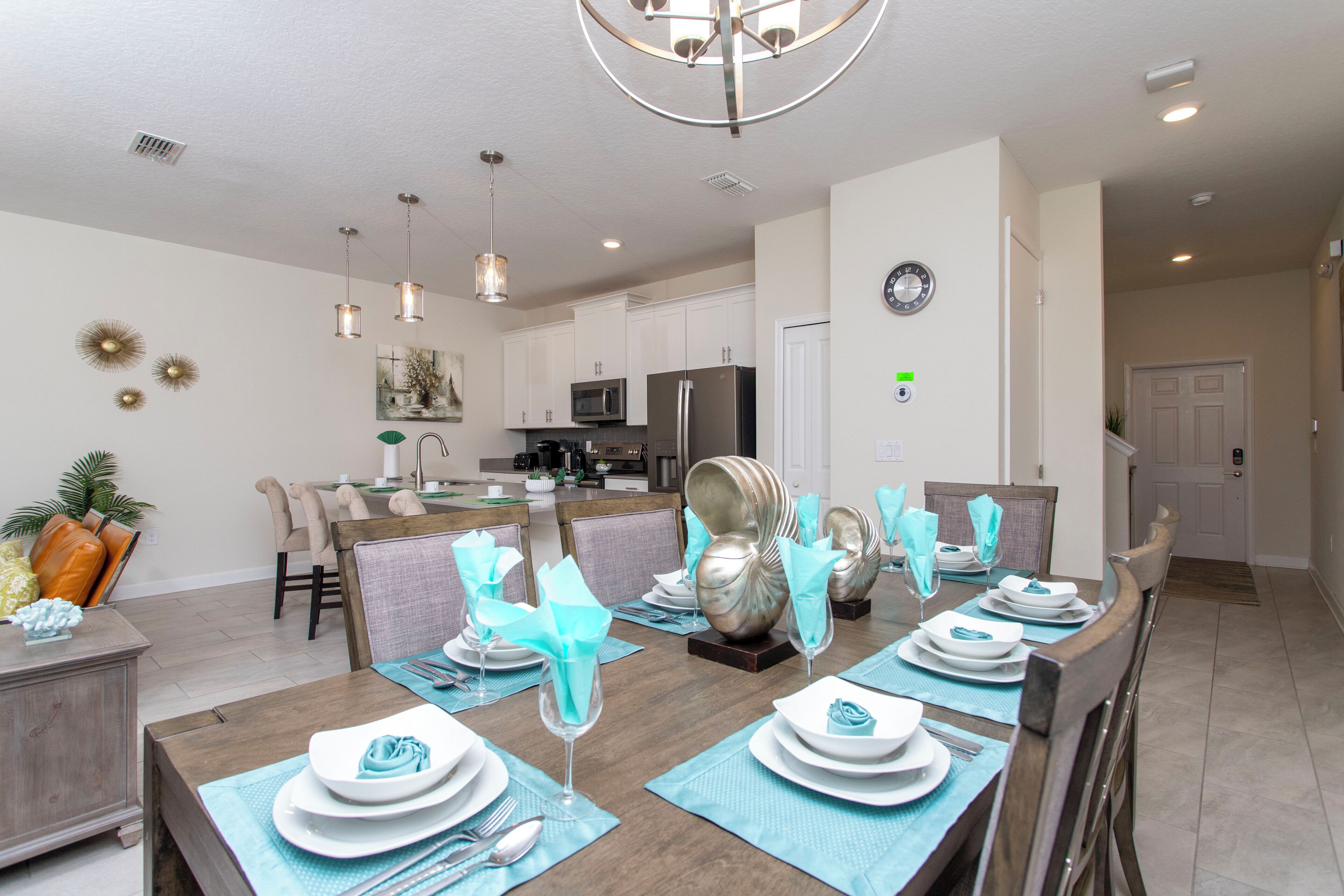 A dining area with a long table set for six, a kitchen in the background, and a clock on the wall.