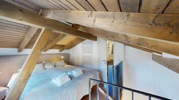 Un dormitorio con techo inclinado, vigas de madera, cama con ropa blanca, lámpara de noche y ventana con persianas.