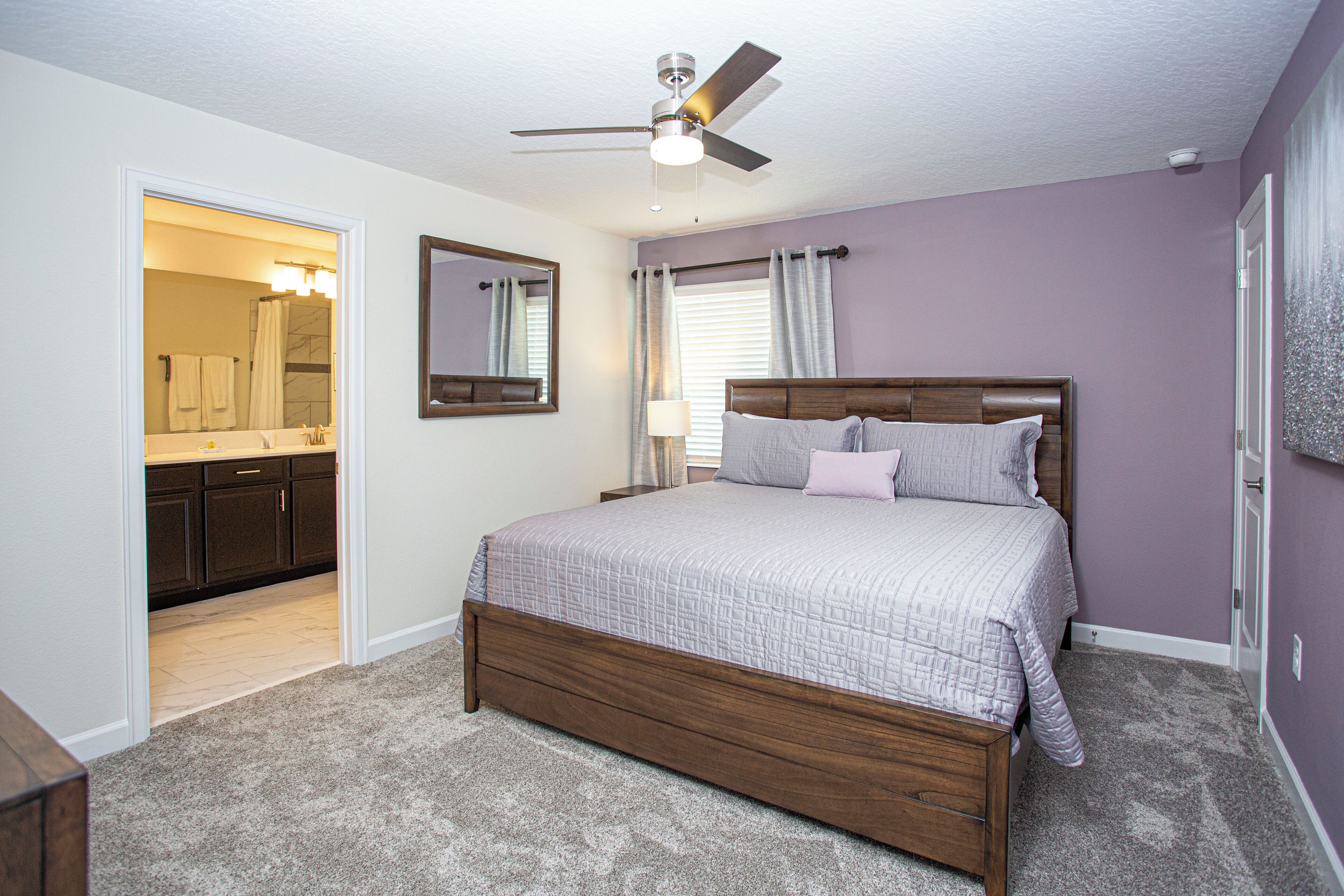 A bedroom with a wooden bed, a ceiling fan, a bathroom with a vanity and mirror, and a view of the kitchen.