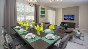 A dining area with a long table set for six, surrounded by chairs. A television is mounted on the wall in the background.