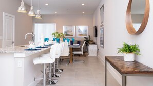 A modern kitchen with white bar stools, pendant lights, and a dining area with a wooden table and chairs. - Gorgeous 4 Bedroom Family Friendly Close to Disney 7573 (Kissimmee)