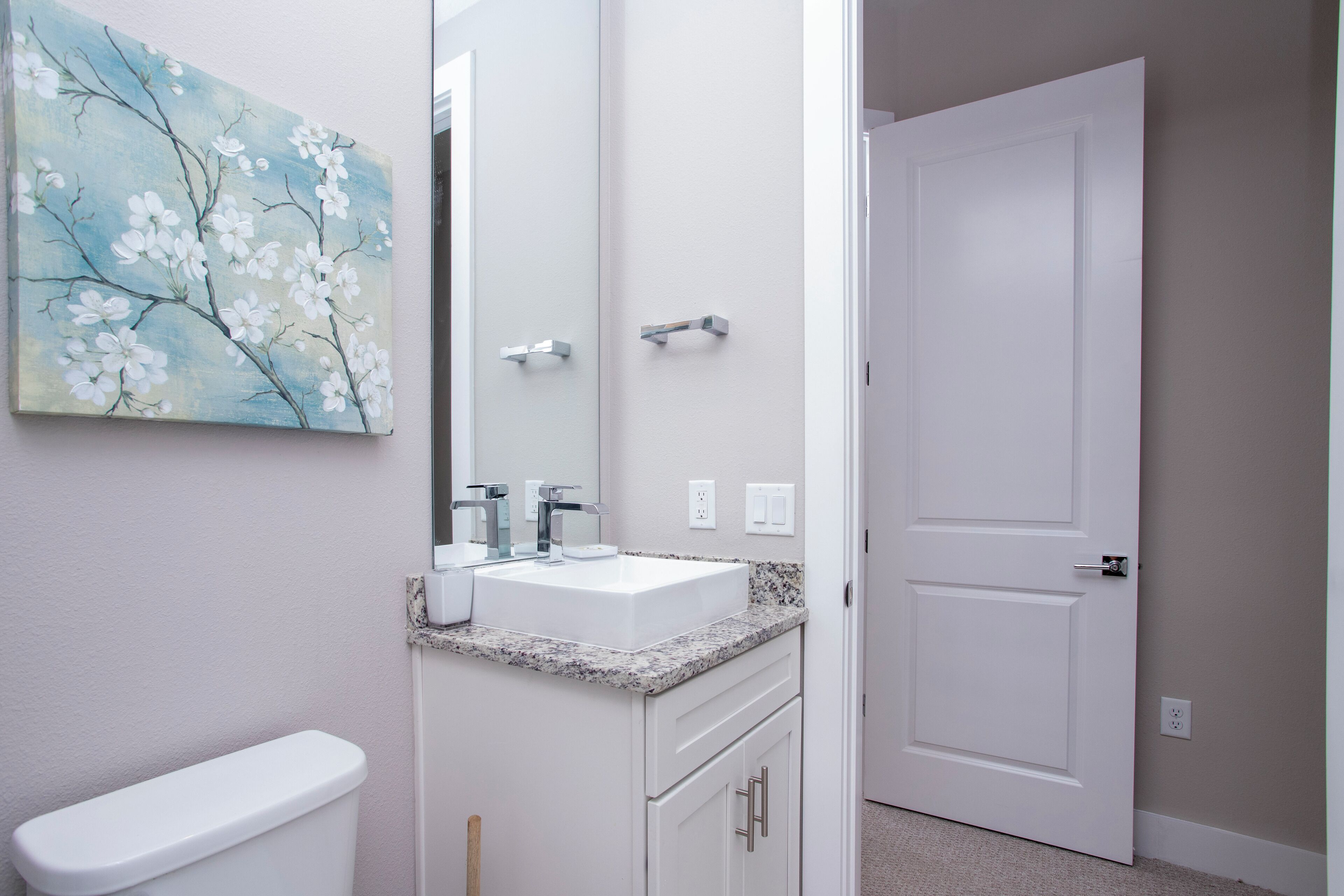 Une salle de bain avec des toilettes blanches, un lavabo doté d’un plan de travail en granit, un miroir et une porte.
