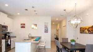 A modern kitchen with white cabinetry, a central island, and a dining area with a dark table and chairs. - Stunning 5 Bd Home w Pool Close to Disney 4822 (Kissimmee)