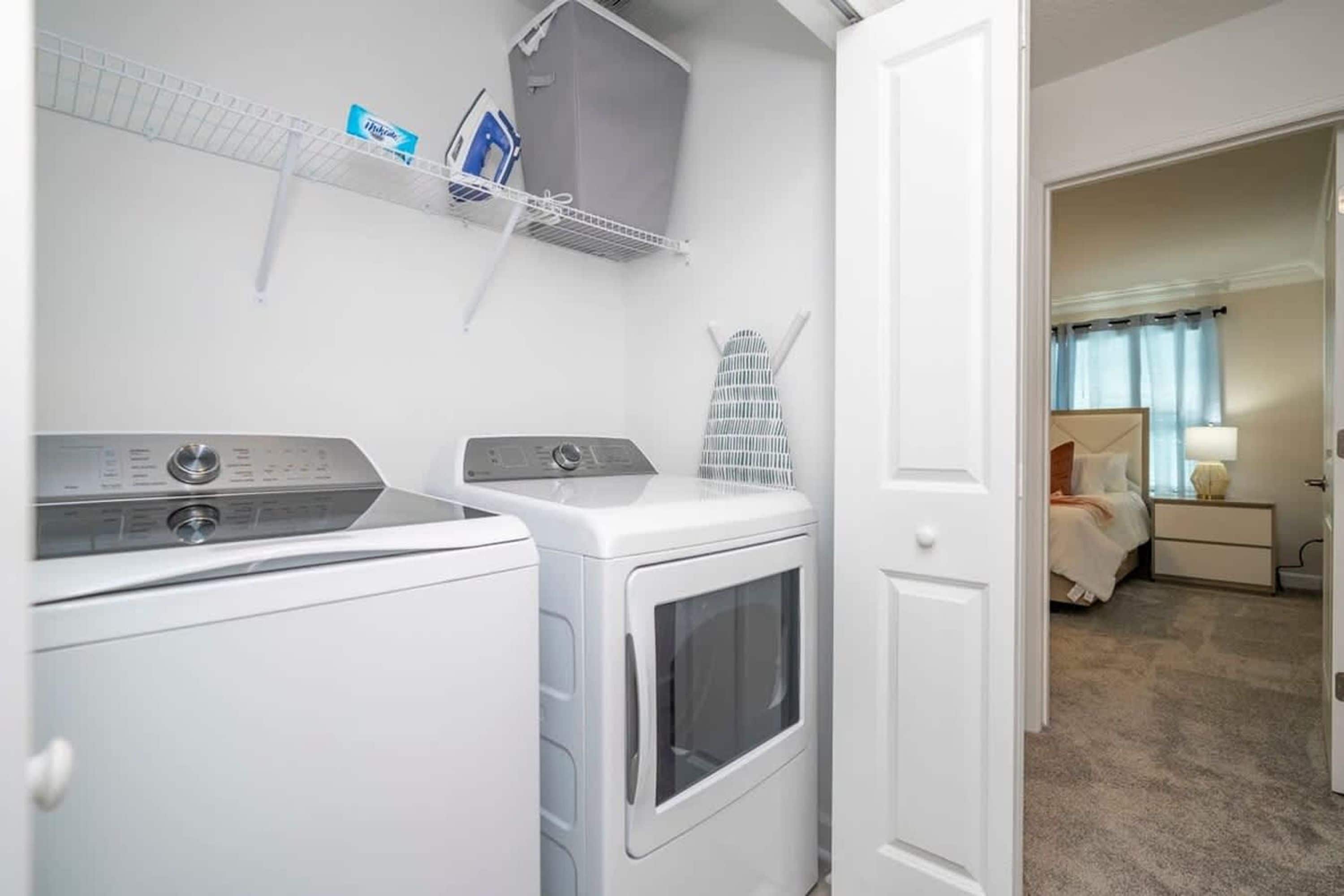 A laundry area with two washing machines and a dryer, a shelf with cleaning supplies, and a bedroom visible through an open door.