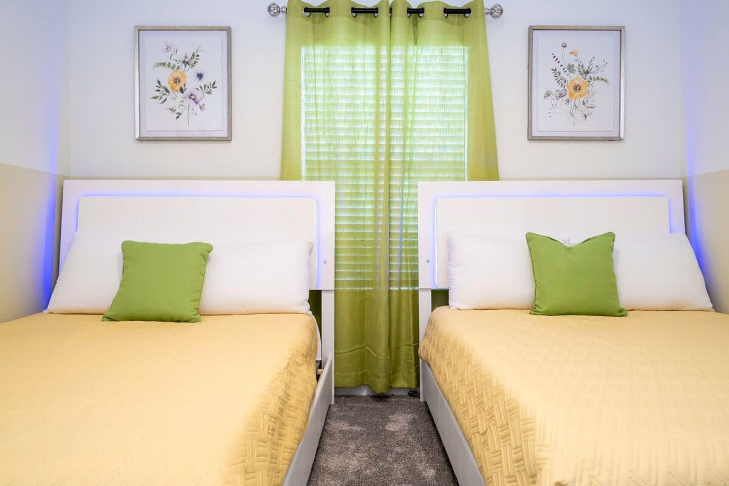 A twin room with two beds, green pillows, a green curtain, and framed floral artwork on the walls.