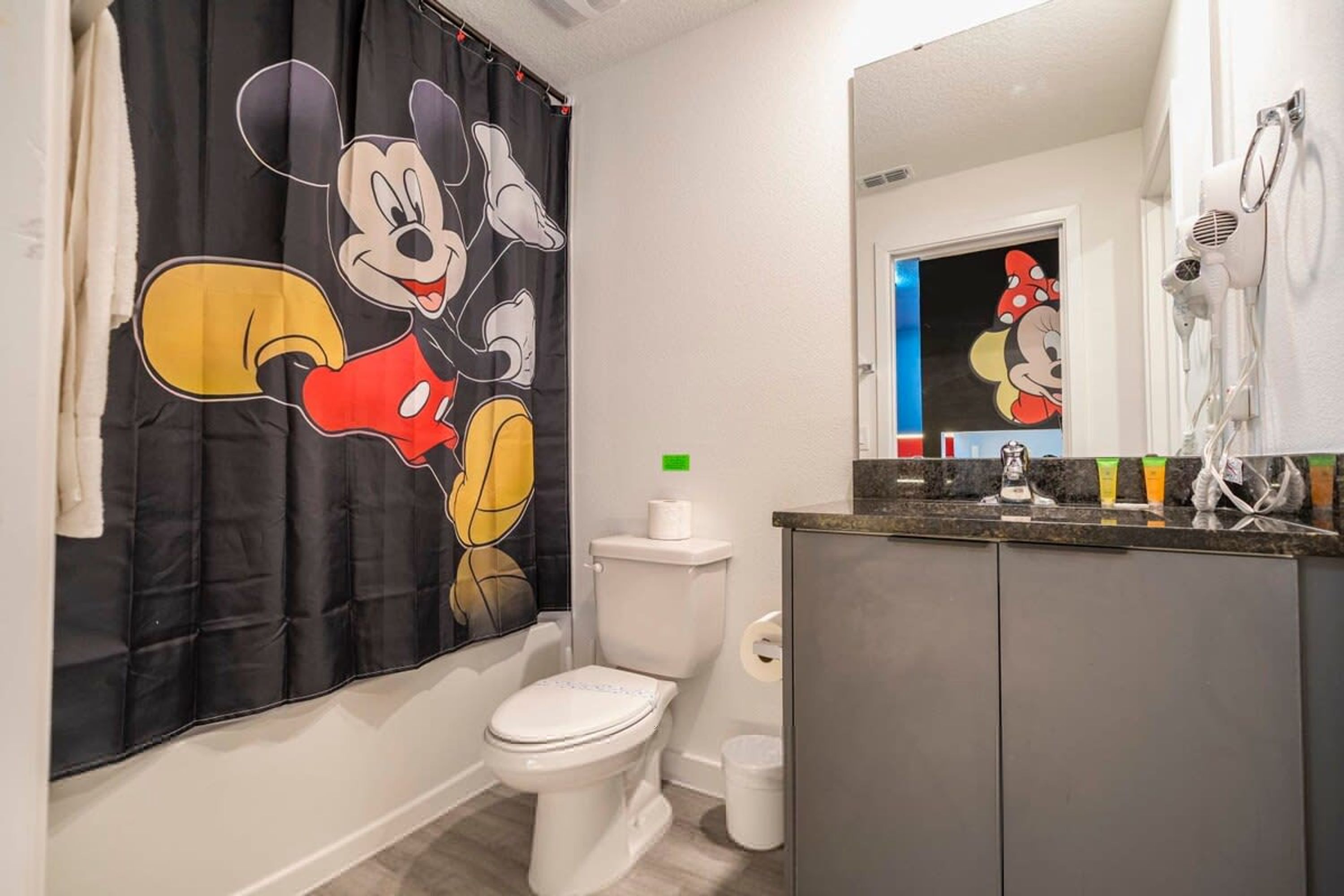Casa de banho com cortina de chuveiro temática do Mickey Mouse, sanita, lavatório com espelho e secador de cabelo na parede.